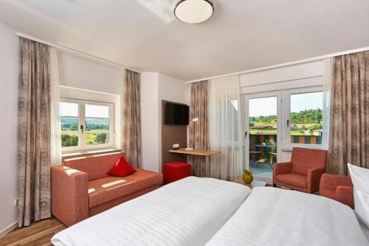 Habitación de hotel con una cama grande, un sofá, una silla y un televisor. Cuenta con un balcón y vistas al campo.