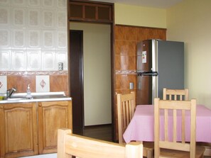 A compact kitchen with wooden cabinets, a stainless steel refrigerator, a sink, and a dining table with a checkered tablecloth. - Portal Austral (Puerto Montt)