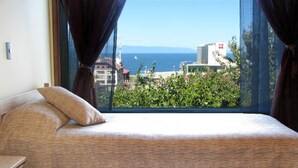 Et soveværelse med en seng, et trænatbord og udsigt til havet og bygninger gennem store vinduer.