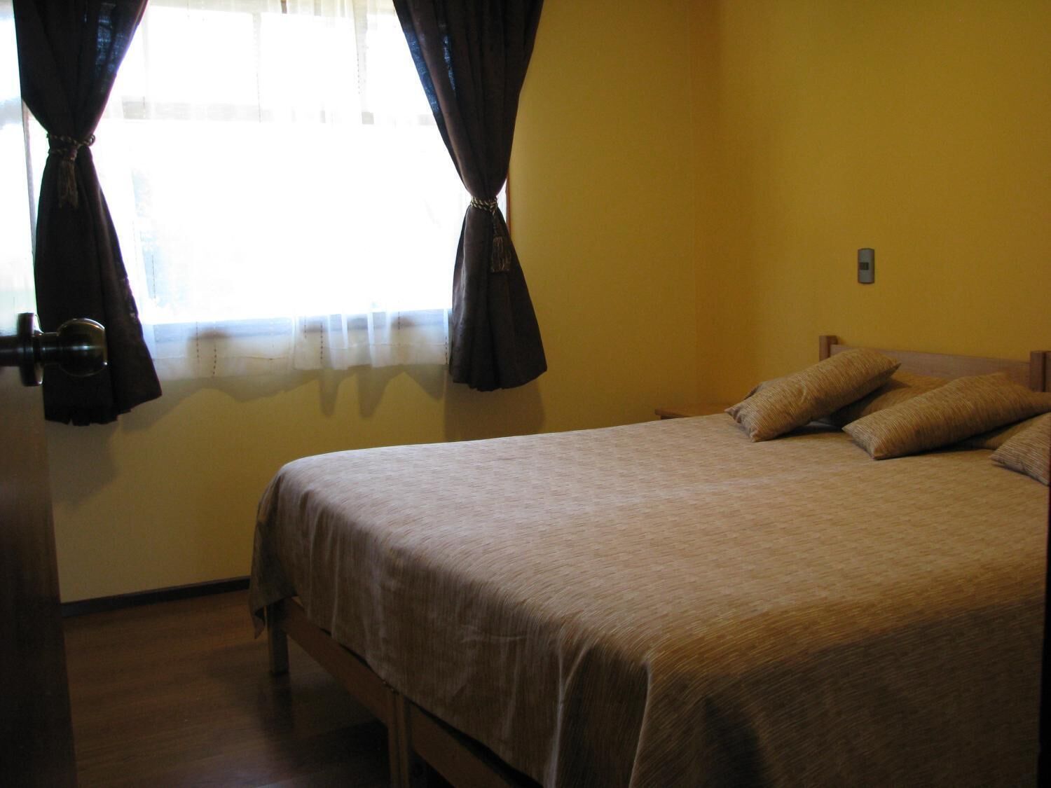 Una cama individual con estructura de madera y colcha color beige, cortinas en una ventana y una pared amarilla lisa.