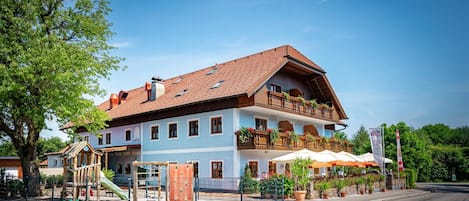 A two-story building with a red-tiled roof, a balcony with plants, and a playground with a slide.