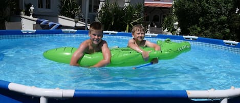 Dos niños en una piscina con flotadores, un borde de piscina azul y un edificio con un letrero 'Zipfer' al fondo.