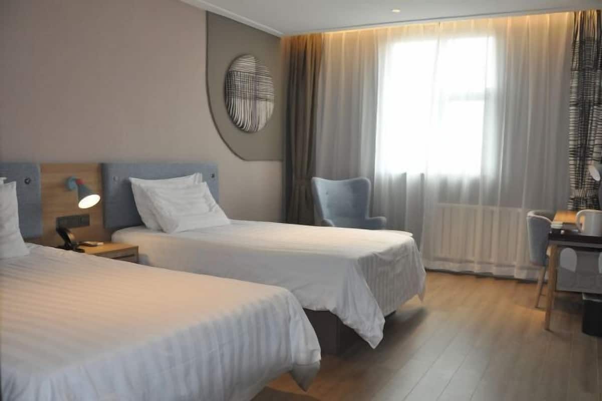 A hotel room with two beds, a desk, a chair, a lamp, and a large window with curtains.