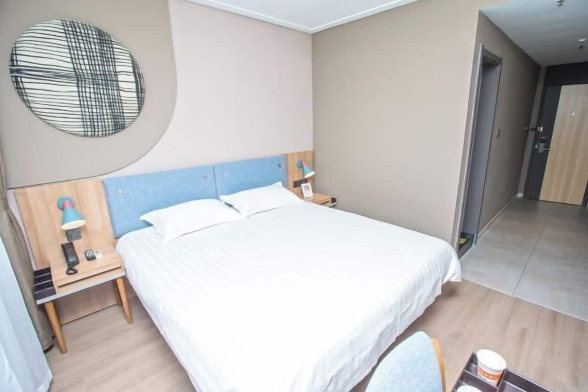 A hotel room with a large bed, a bedside table with a lamp, a round wall decoration, and a door leading to another room.