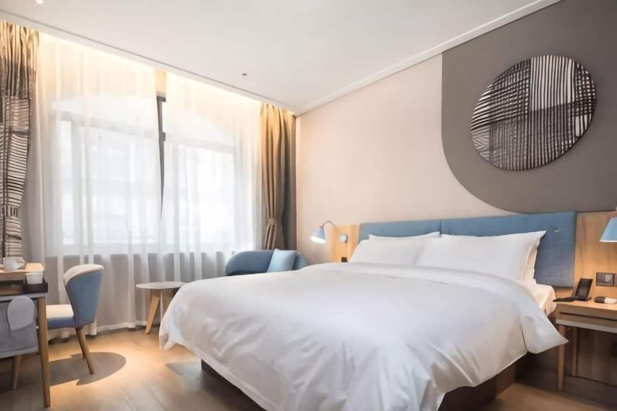 A hotel room with a large bed, a desk with a chair, a small table, and a round wall decoration.