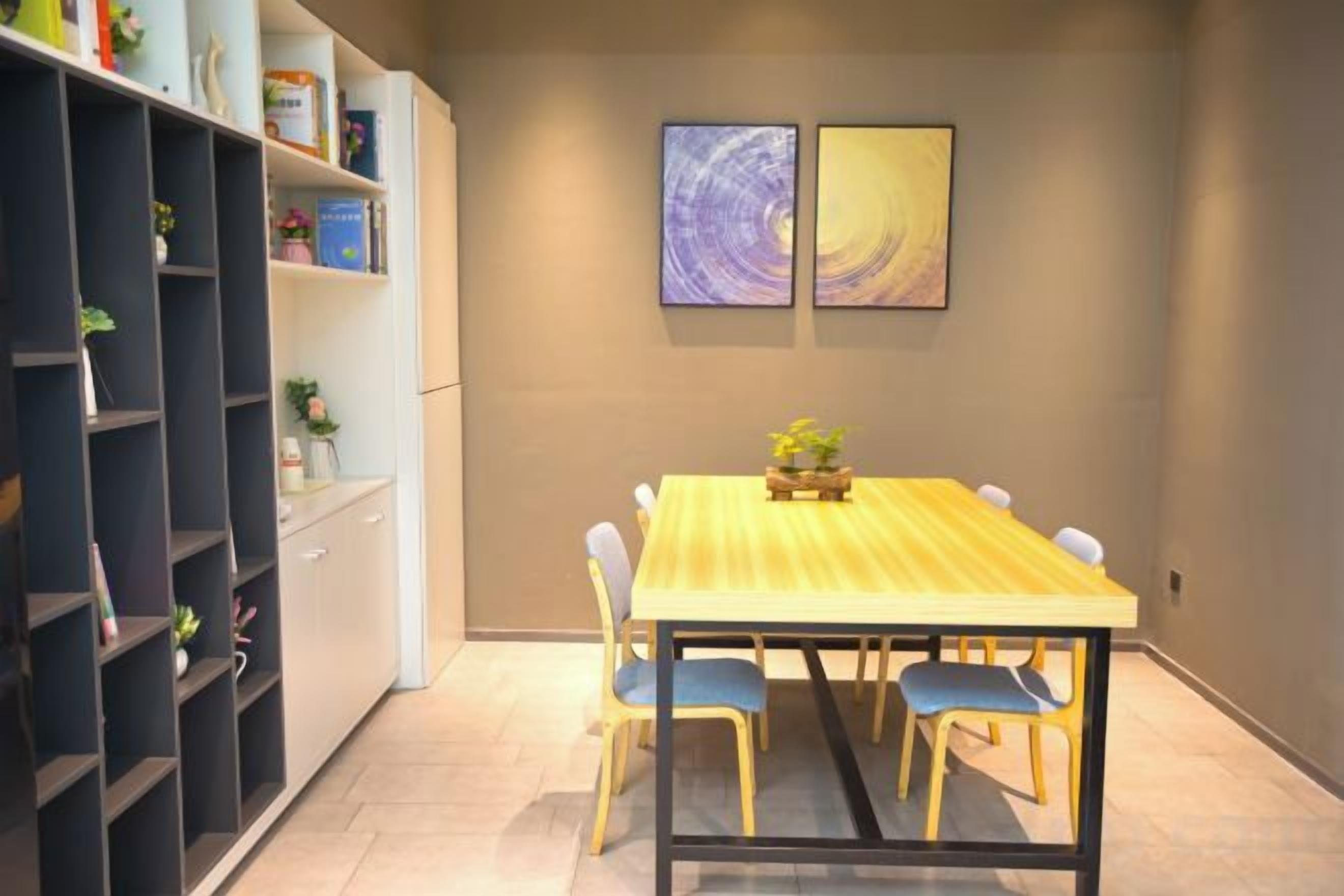 A dining area with a wooden table and chairs, a bookshelf, and two framed artworks on the wall.