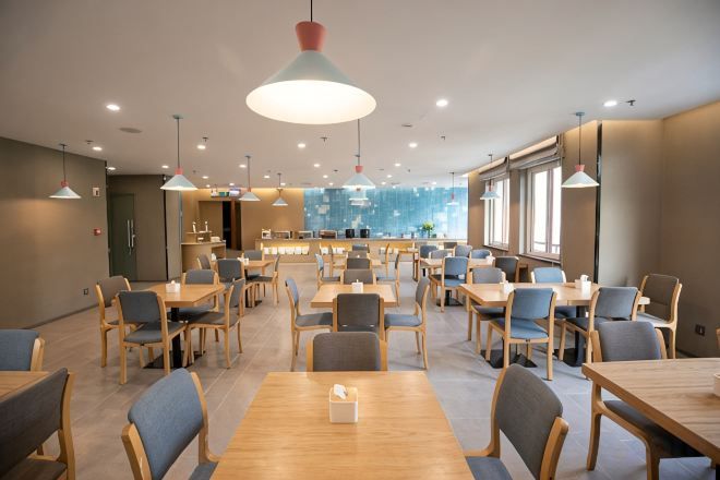Amplia zona de comedor con varias mesas y sillas, un bar y grandes ventanales que permiten la entrada de luz natural.