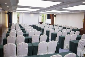 Un salón de conferencias con filas de mesas y sillas, una pantalla de proyector y un atril para presentaciones.