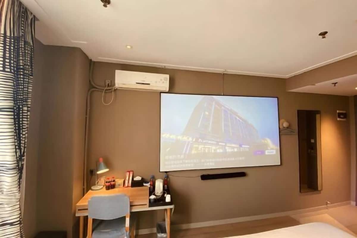 A hotel room with a bed, a desk with a chair, a television mounted on the wall, and an air conditioning unit.