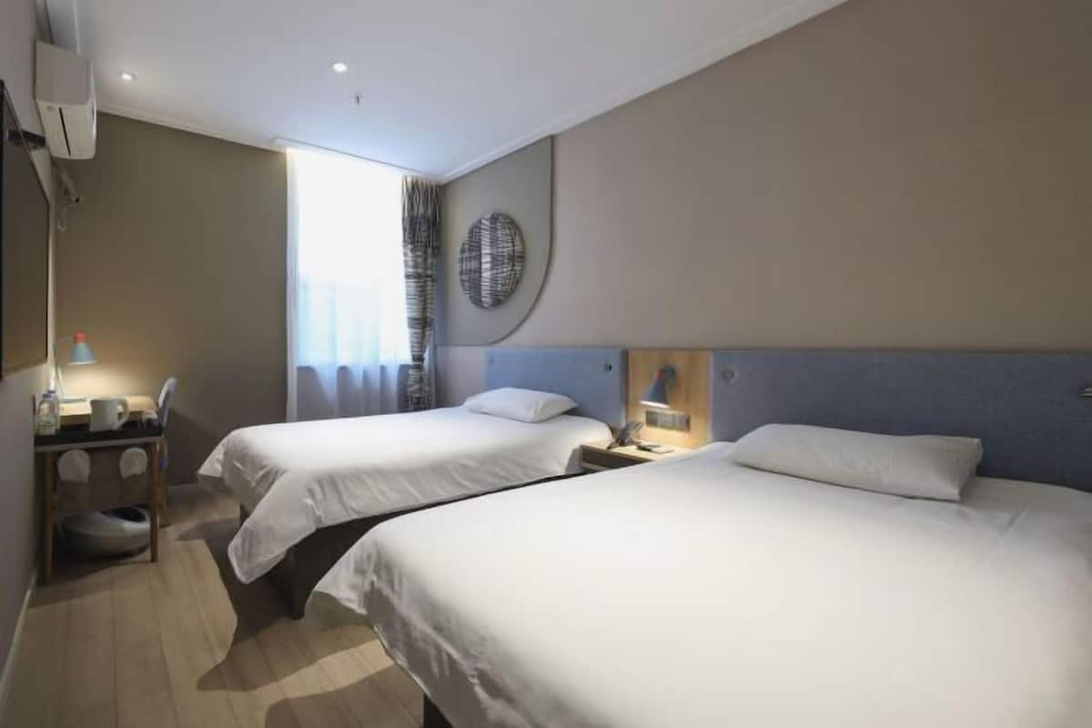 A hotel room with two beds, a desk with a chair, a lamp, and a large window with curtains.