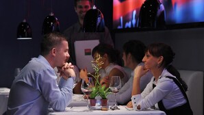 Eine Gruppe von Personen sitzt an einem Tisch, Wein gläser und eine Pflanze stehen bereit. Der Raum ist schummrig beleuchtet, im Hintergrund ist ein großer Bildschirm zu sehen.