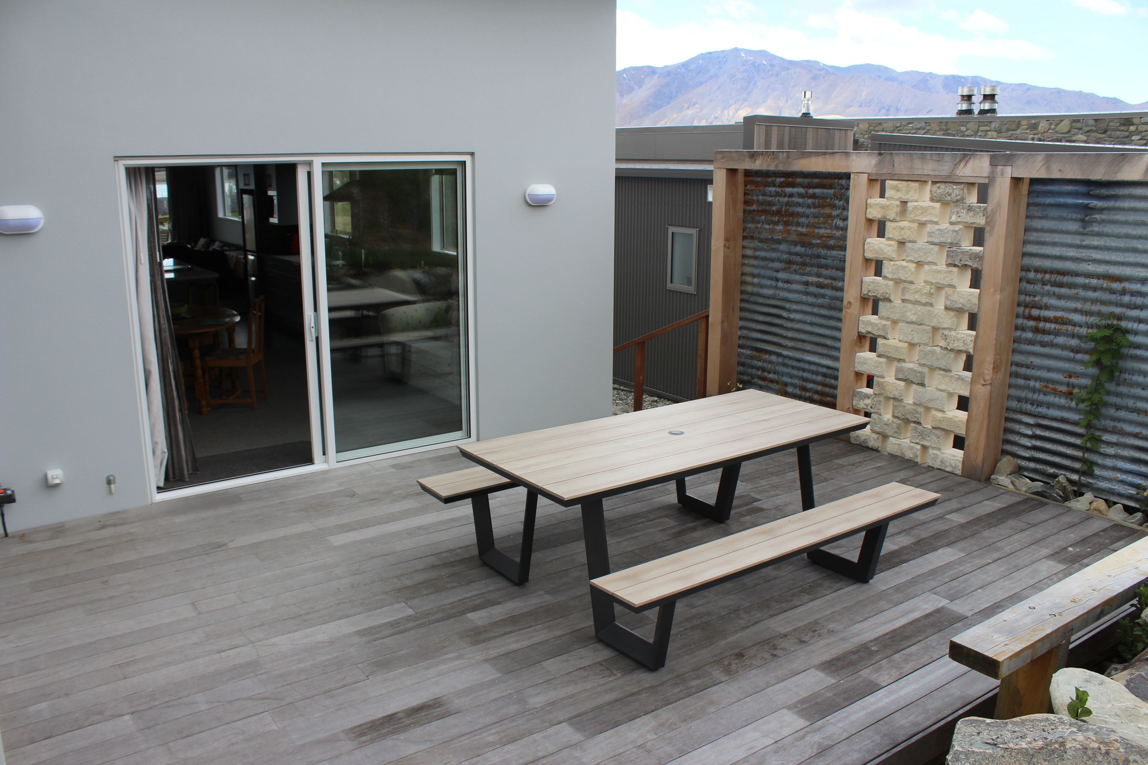 Un patio al aire libre con una mesa de madera y bancos, un cerramiento de vidrio corredizo que da a una habitación, y una pared de metal corrugado con detalles de piedra.
