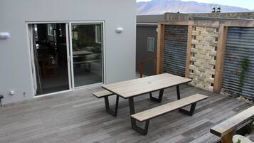 Un patio al aire libre con una mesa de madera y bancos, un cerramiento de vidrio corredizo que da a una habitación, y una pared de metal corrugado con detalles de piedra.