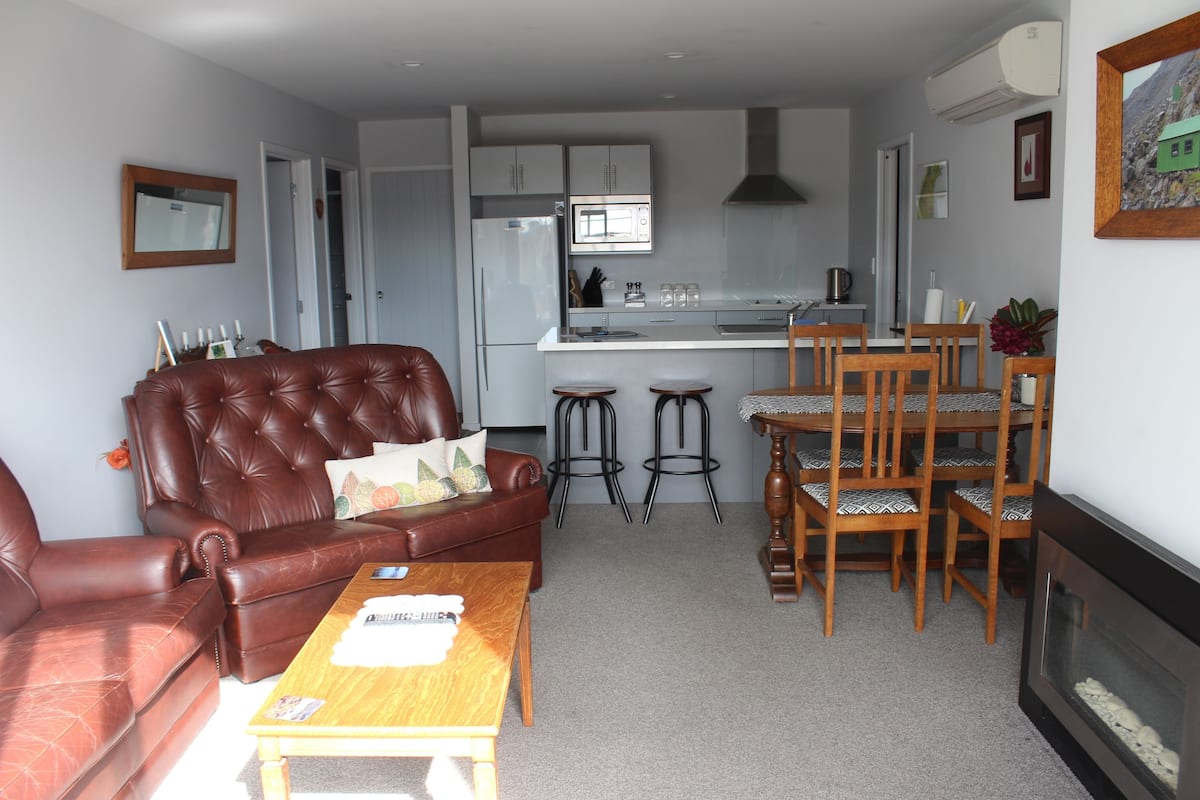 A living room with a brown leather sofa, a wooden coffee table, and a fireplace. There is a kitchen area with a microwave, refrigerator, and bar stools.