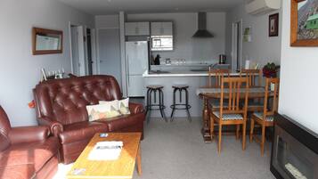 Een woonkamer met een bruine leren bank, een houten salontafel en een open haard. Er is een keukenhoek met een magnetron, een koelkast en barstoelen.