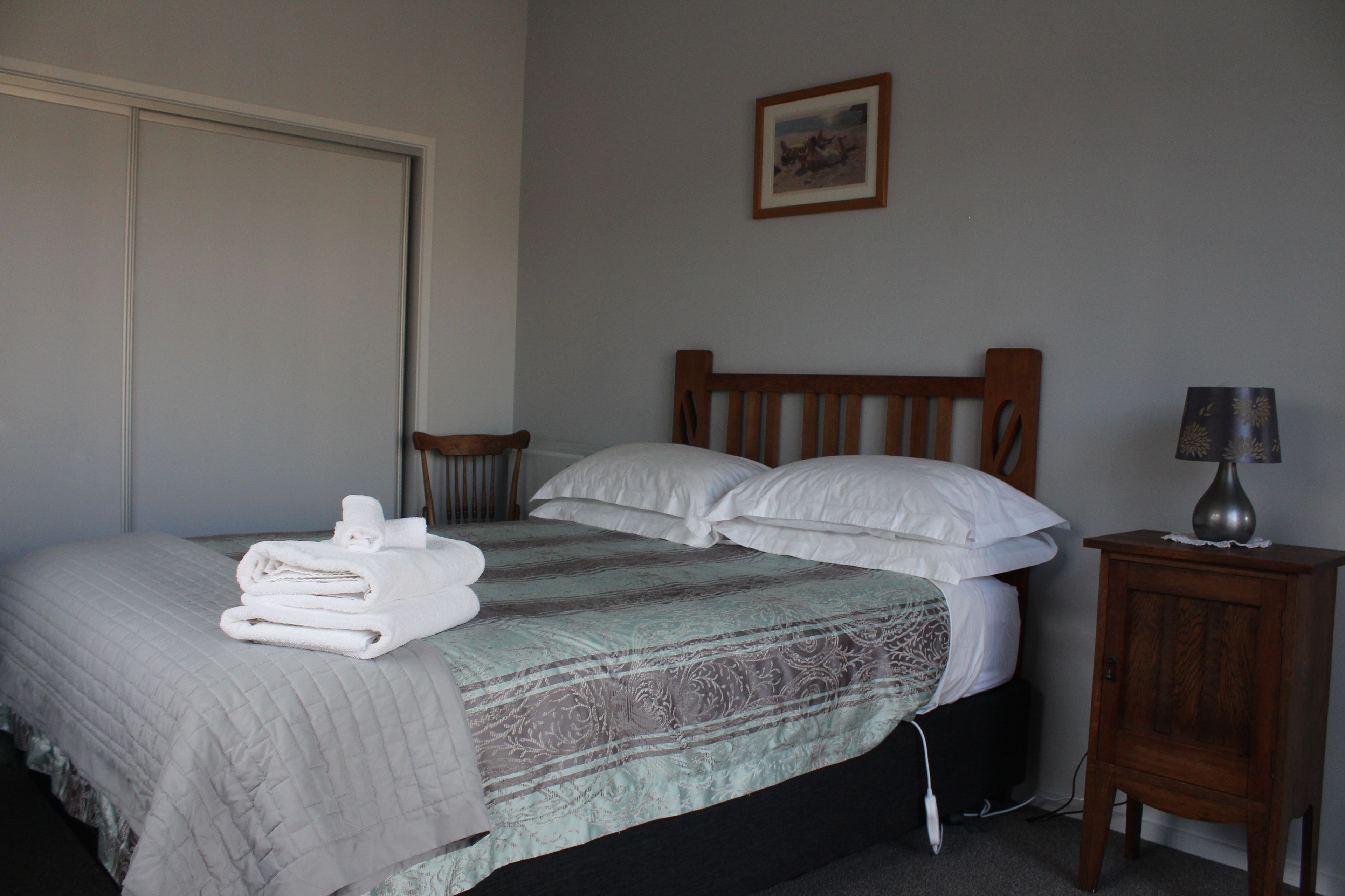 A neatly made bed with a quilt, pillows, and folded towels. There is a small wooden nightstand with a lamp beside the bed.