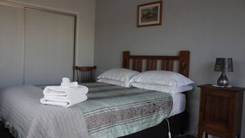 A neatly made bed with a quilt, pillows, and folded towels. There is a small wooden nightstand with a lamp beside the bed.