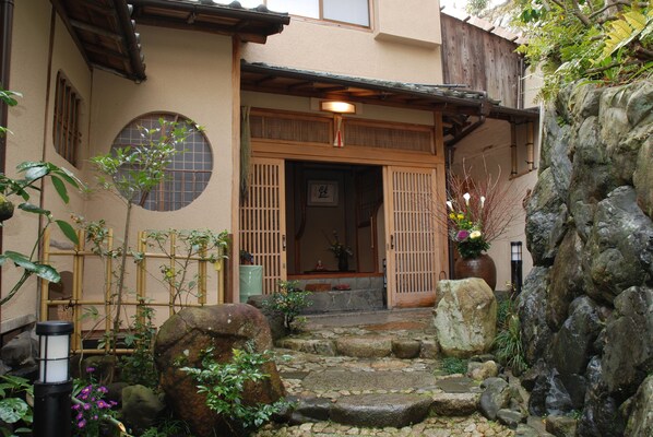 A traditional Japanese building with a wooden entrance, stone steps, and a garden with rocks and plants.