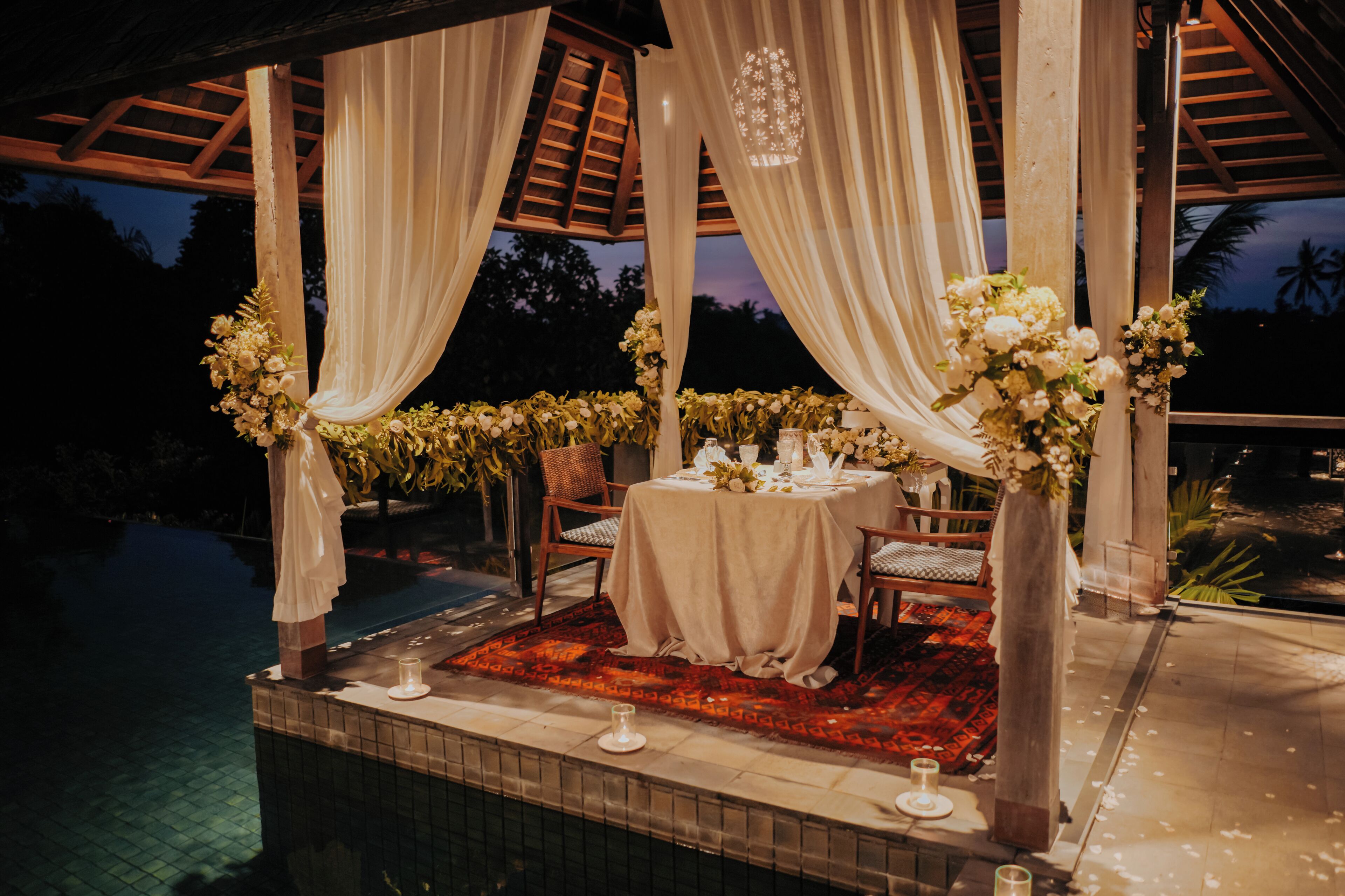 An outdoor dining area with a pool, draped white curtains, and floral centerpieces.