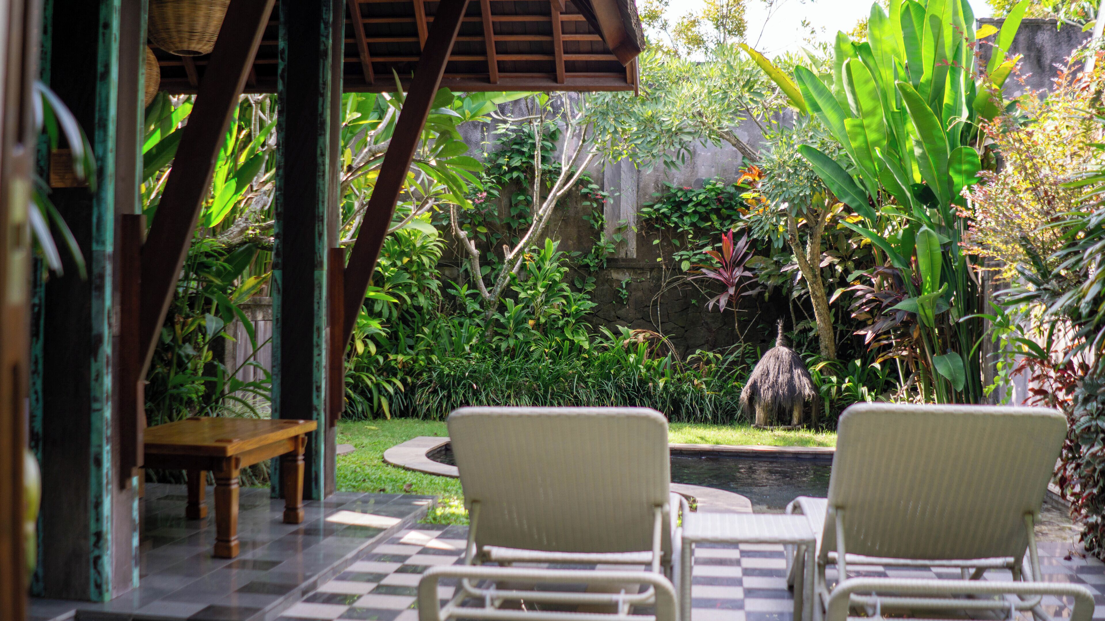 A patio with two white wicker chairs, a wooden table, and a small pond in a lush garden.