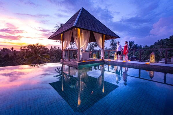 A pool with a gazebo, palm trees, and a sunset.