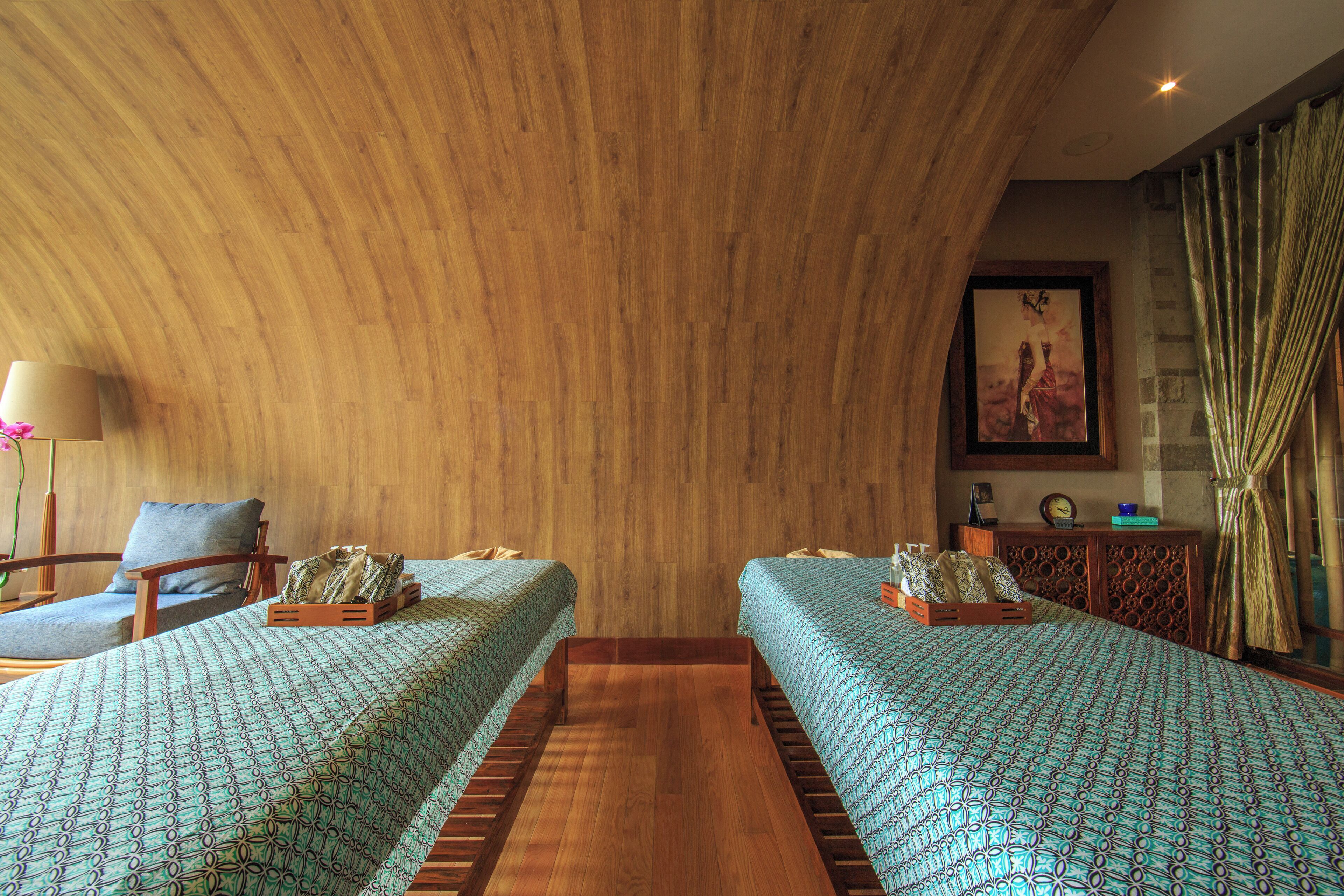 Una habitación con dos camas, suelo de madera, una silla, una lámpara y un cuadro en la pared.