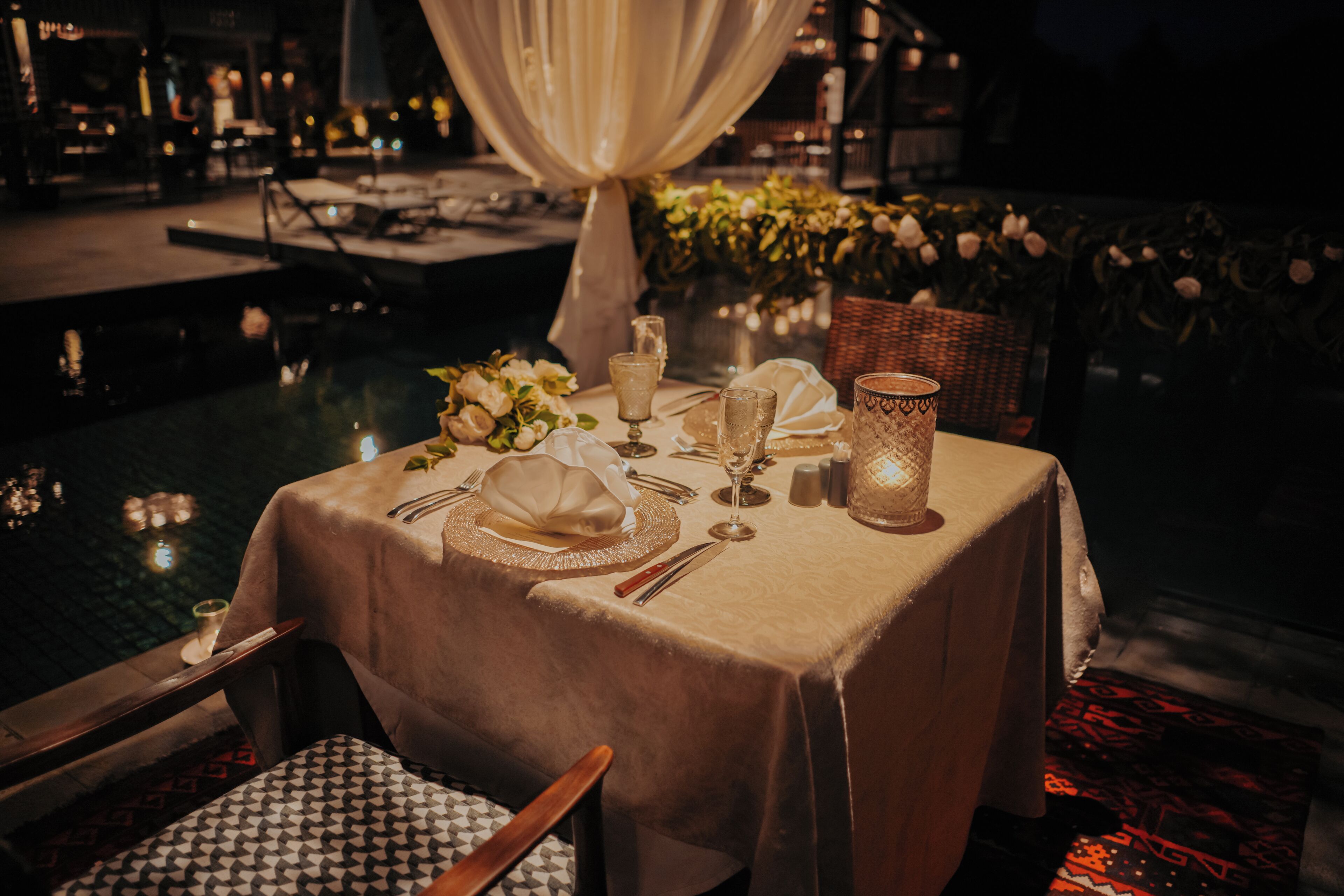 Mesa lista para dos con vista a una piscina, una vela y un jarrón de flores.