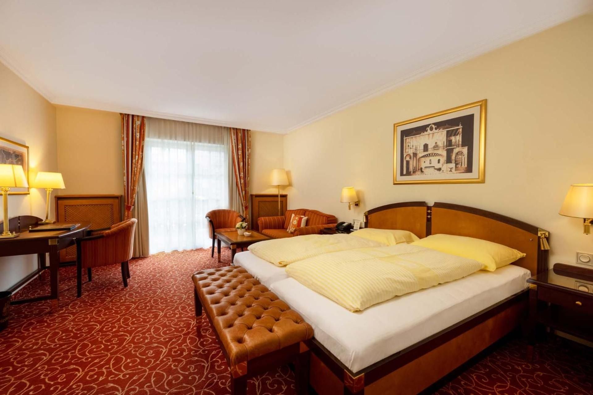 A hotel room with a bed, a desk, a chair, a bench, and a painting on the wall.