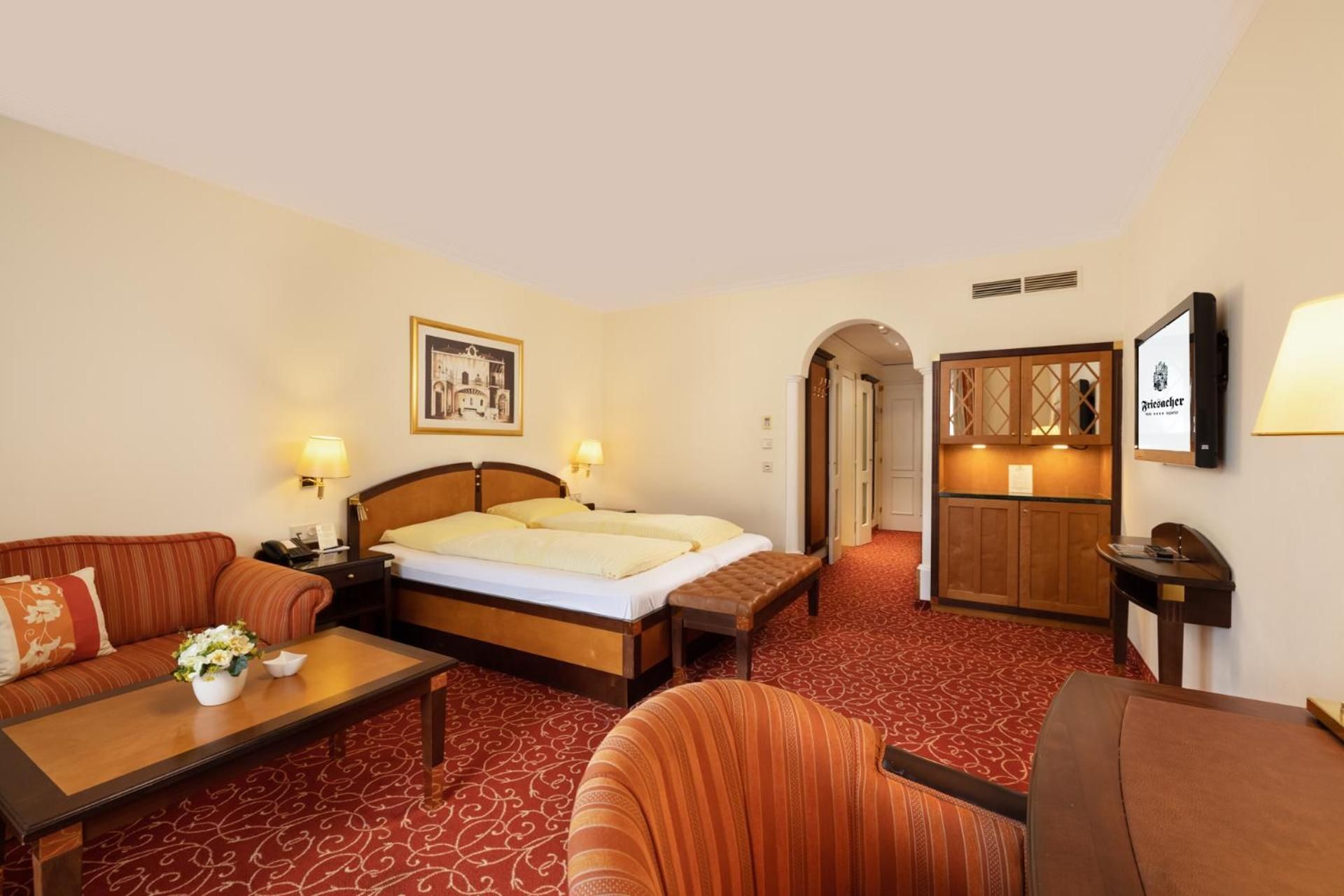 A hotel room with a bed, a sofa, a small table with a vase of flowers, a desk, and a cabinet.