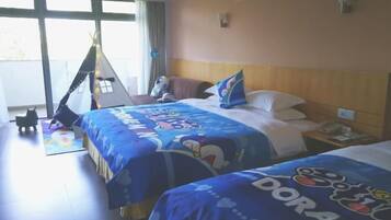 A hotel room with two beds, each with a blue bedspread featuring cartoon characters. There is a window with curtains, a nightstand with a phone, and a small table with a chair.