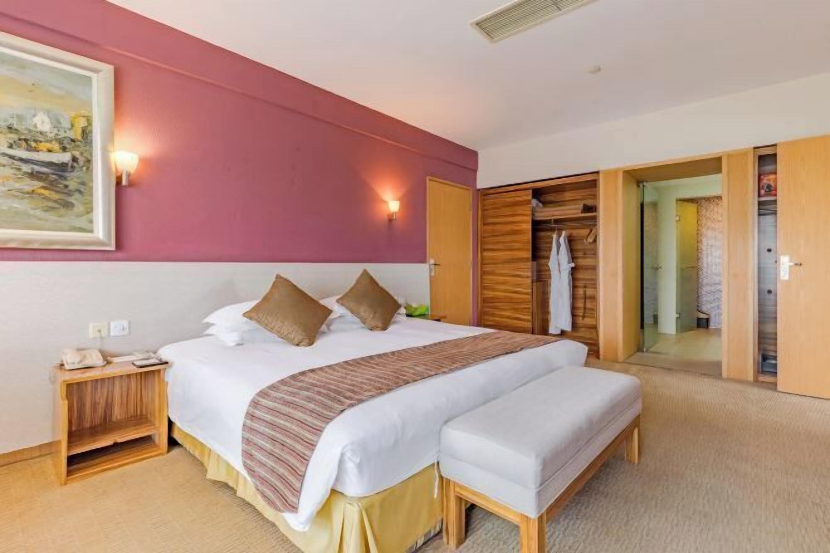 Ett hotellrum med en säng, sängbord, en bänk, en garderob och en tavla på väggen.