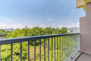 Un balcon doté d’une balustrade en métal, donnant sur un paysage verdoyant avec des arbres et des bâtiments situés au loin.