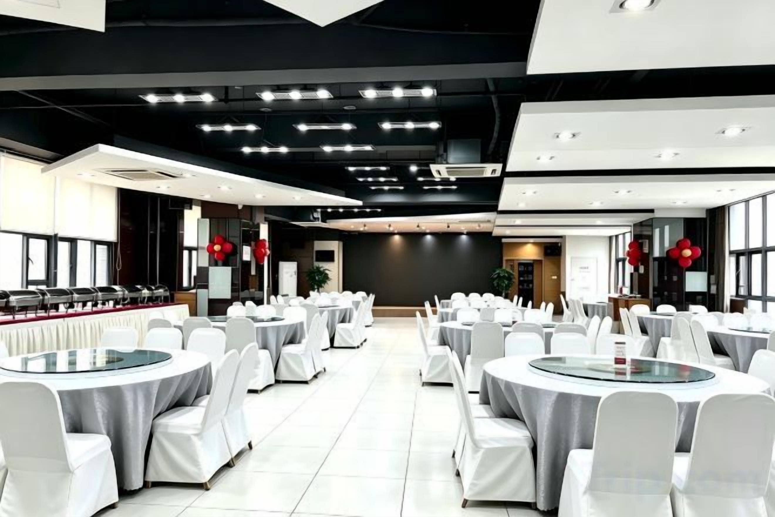 Un salón de banquetes con mesas y sillas redondas, una zona de cocina y un diseño de techo moderno.