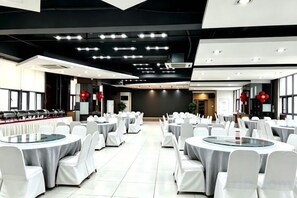 A banquet hall with round tables and chairs, a kitchen area, and a modern ceiling design.