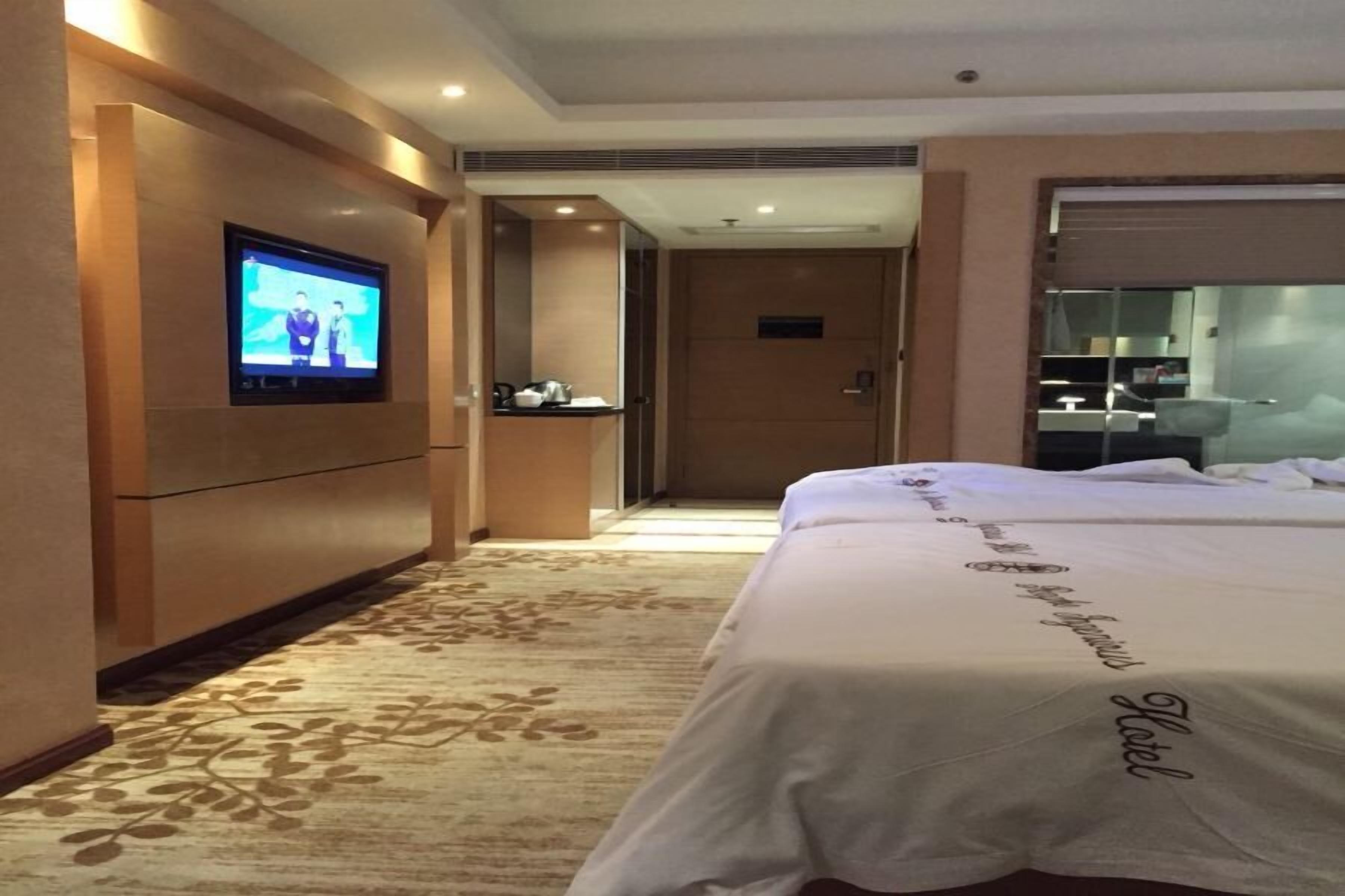 A hotel room with a TV on the wall, a bed with a hotel name embroidered on the bedspread, and a carpet with a floral pattern.