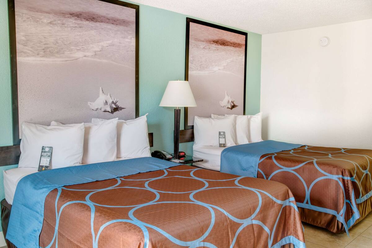A hotel room with two beds, each with a brown and blue patterned bedspread, a nightstand with a lamp, and a framed picture of a beach scene on the wall.
