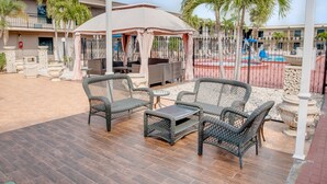 An outdoor patio with wicker furniture and a table, surrounded by palm trees and a pool area.