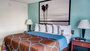 A hotel room with a large bed, two bedside tables with lamps, a nightstand, and a wall-mounted painting of palm trees.