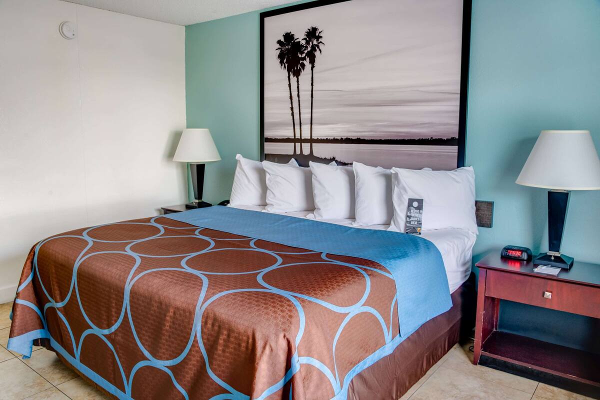 A hotel room with a large bed, two bedside tables with lamps, a nightstand, and a wall-mounted painting of palm trees.