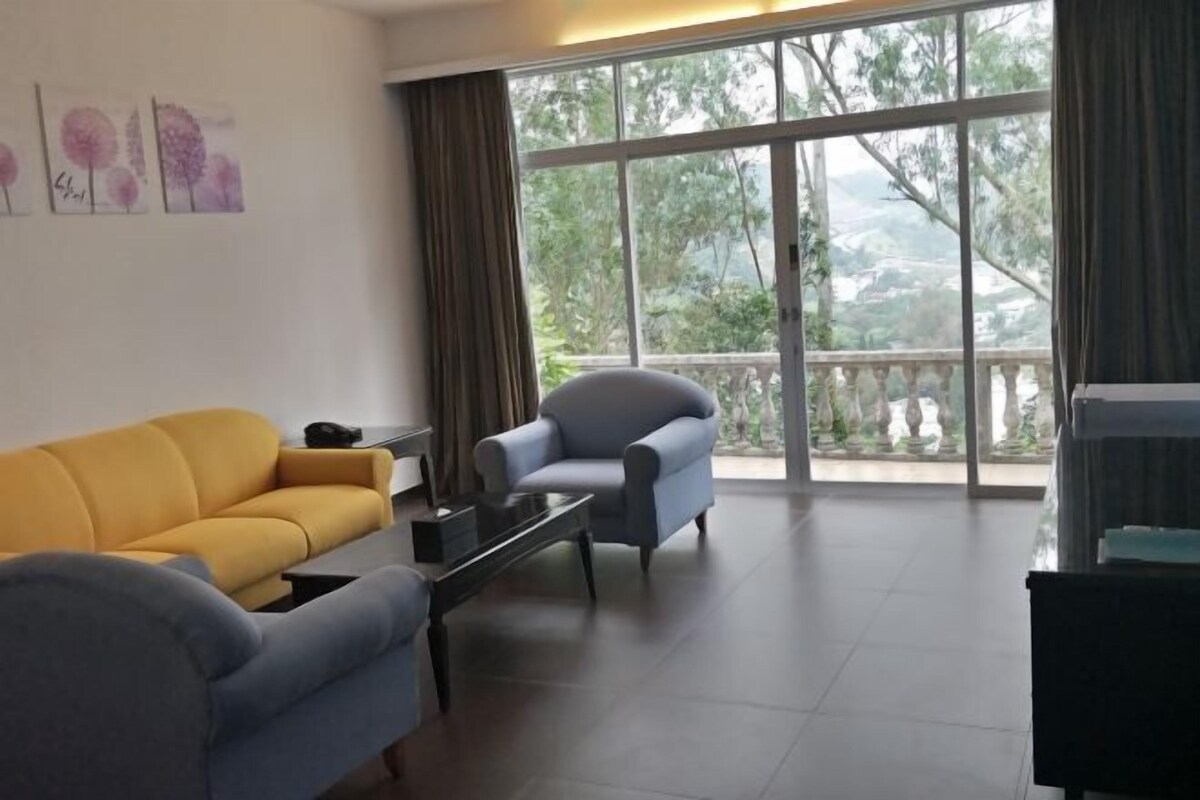 Ein Wohnzimmer mit einem gelben Sofa, einem grauen Sessel und einem schwarzen Couchtisch. Große Fenster bieten Blick auf Grünflächen und einen Balkon.