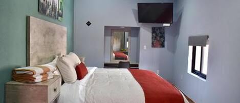 Un dormitorio con una cama, una mesita de noche, un televisor montado en la pared y una ventana.