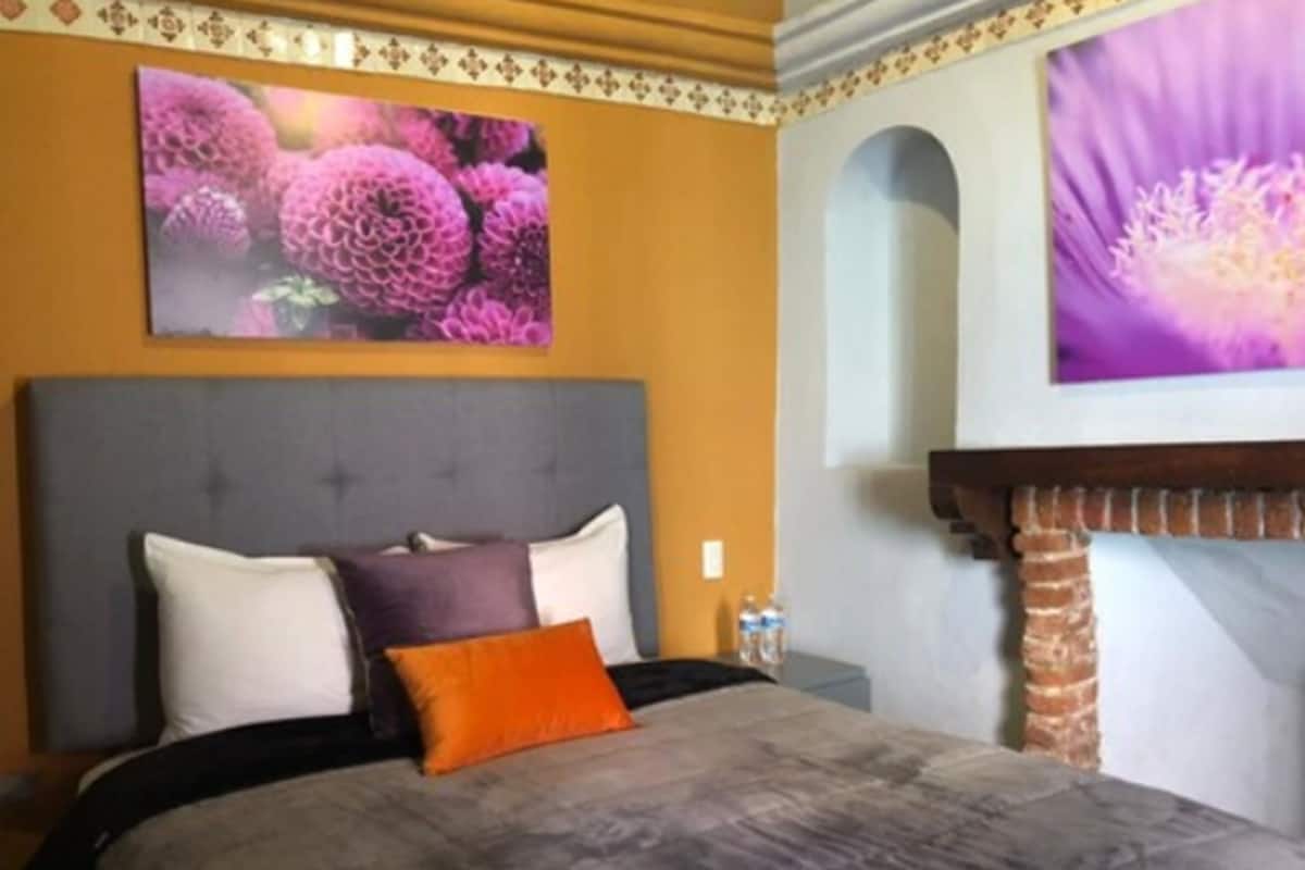 Un dormitorio con cabecera tapizada en gris, cama con edredón gris y naranja, y dos almohadas. Hay un televisor montado en la pared sobre la cama mostrando una imagen floral.