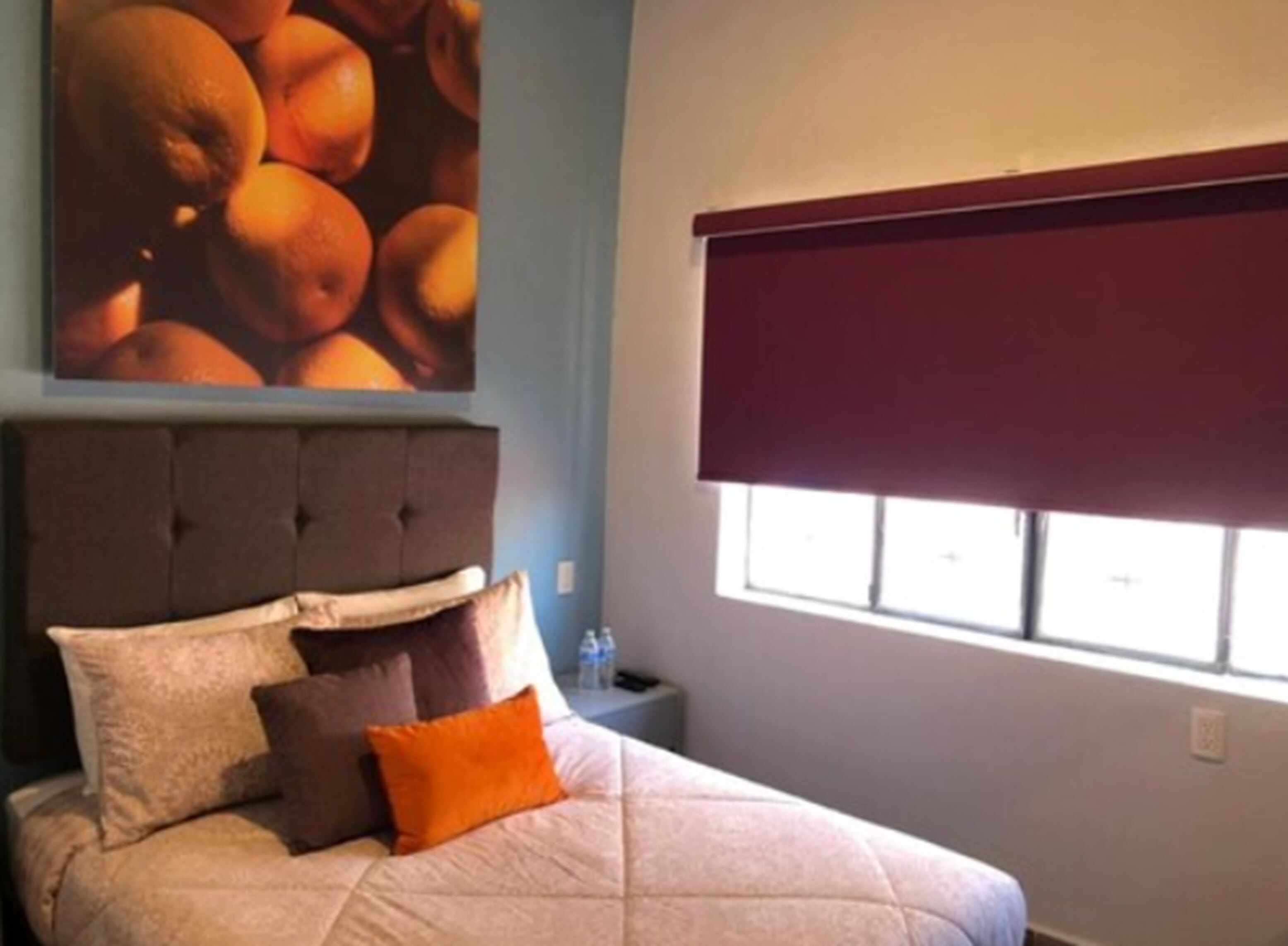 A bedroom with a bed, pillows, a wall-mounted artwork of oranges, a window with a blind, and a nightstand with a bottle.