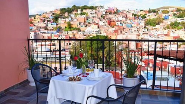 Un balcón con mesa lista para comer, con vista a un pueblo en la ladera lleno de construcciones.