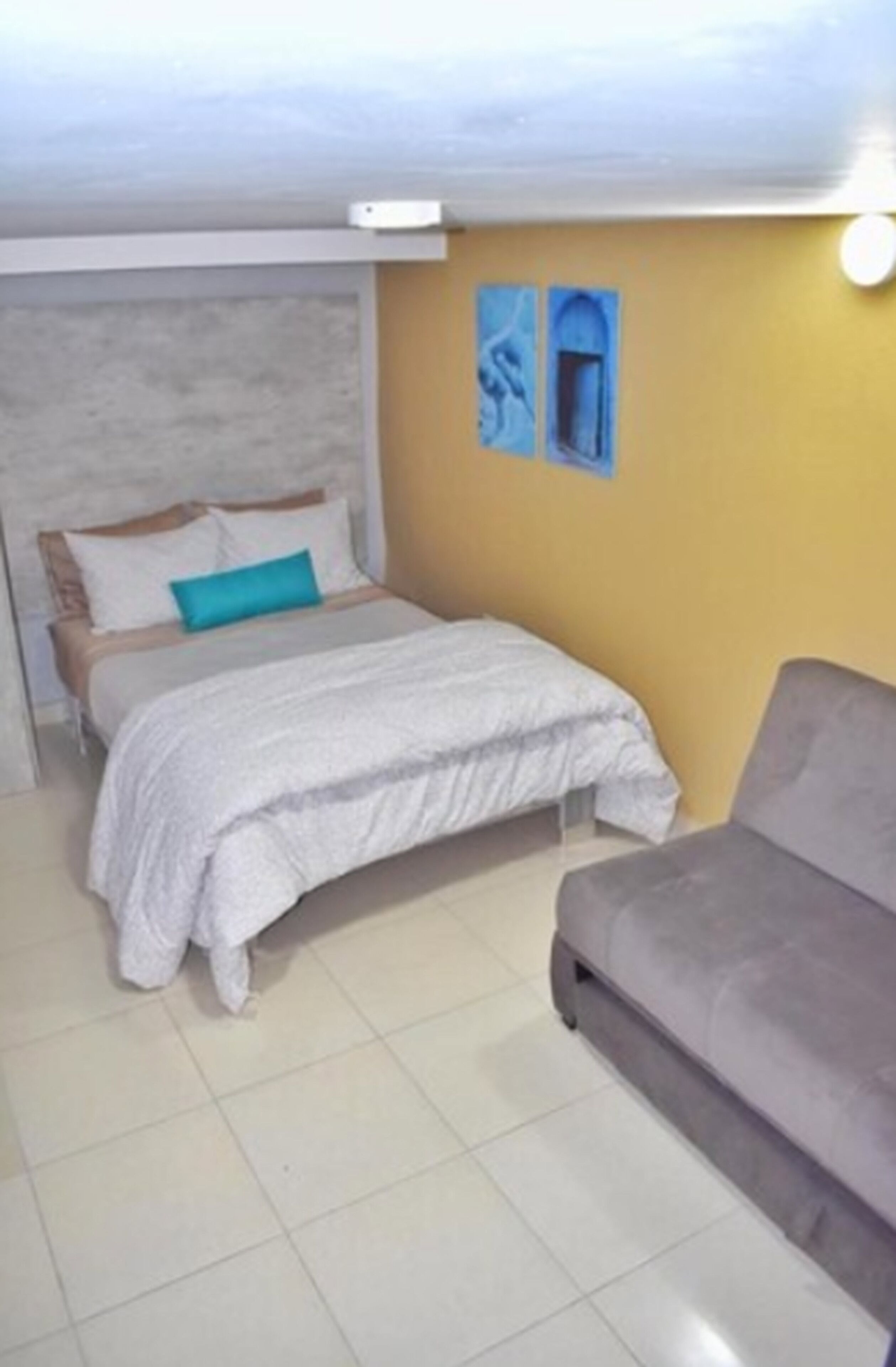 Quarto com uma cama, um sofá cinzento e uma parede amarela.