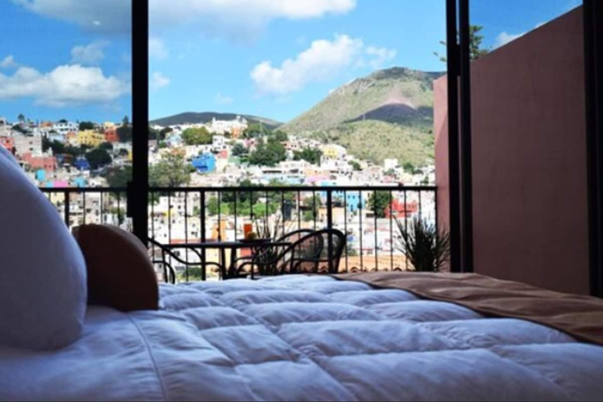 Habitación de hotel con cama, almohadas y vista a un pueblo colorido y colinas.