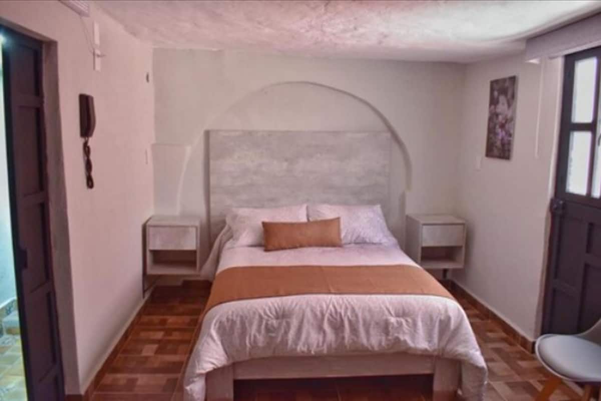 Un dormitorio con cama, mesitas de noche, una silla y una puerta.