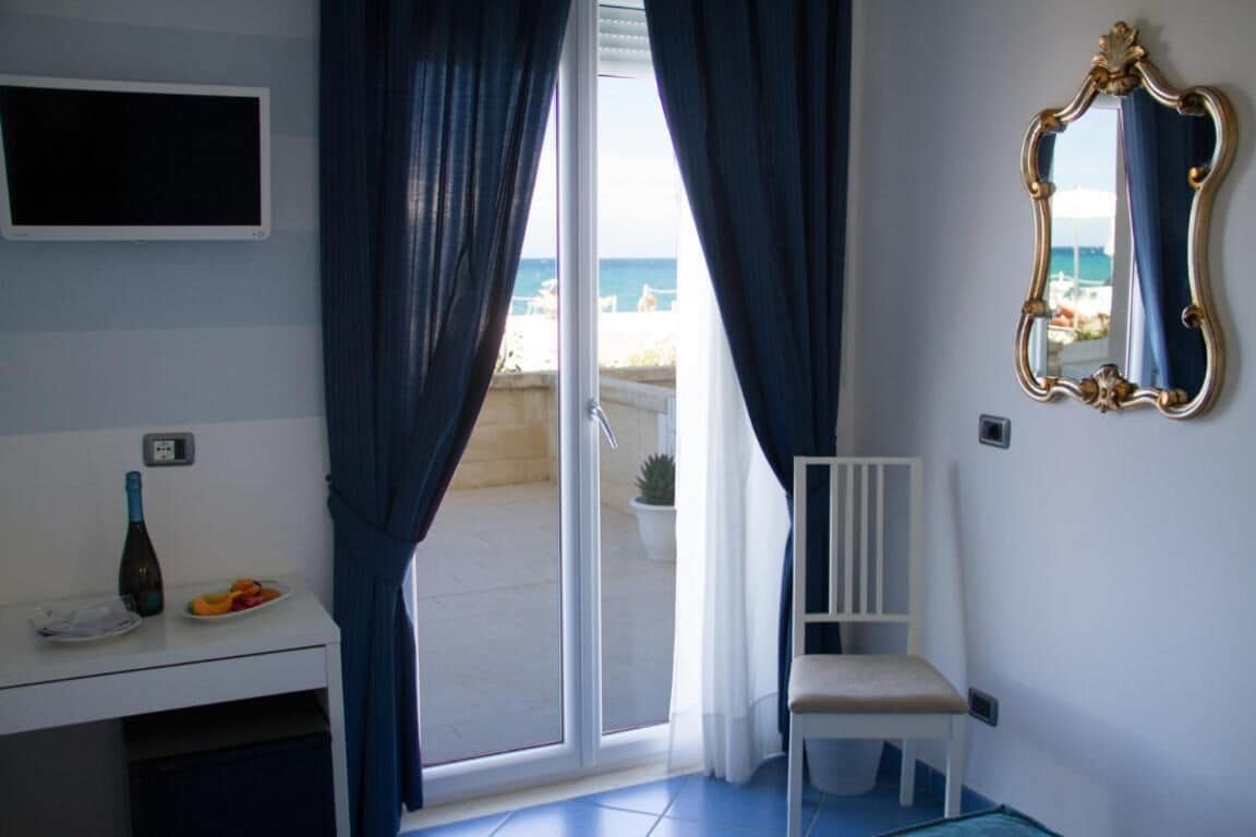 Una stanza con una scrivania bianca, una sedia, uno specchio e vista sulla spiaggia tramite una porta-finestra.
