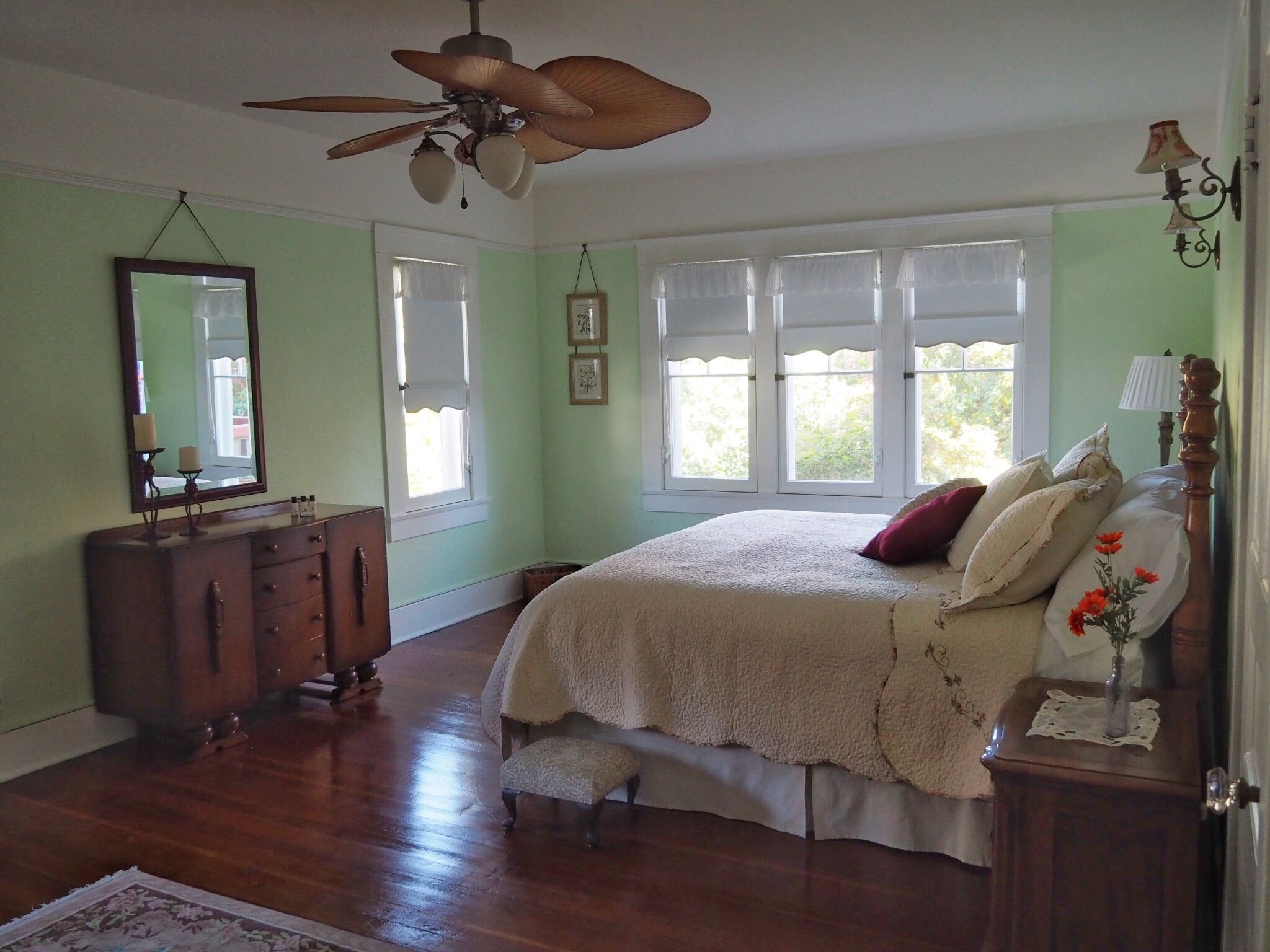 A bedroom with a large bed, a wooden dresser, a ceiling fan, and a window with white curtains.