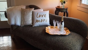 Un sofá estampado de leopardo con una manta blanca esponjosa, un cojín que dice 'Love more', una mesita con una lámpara y flores, y una bandeja con vasos y una botella.
